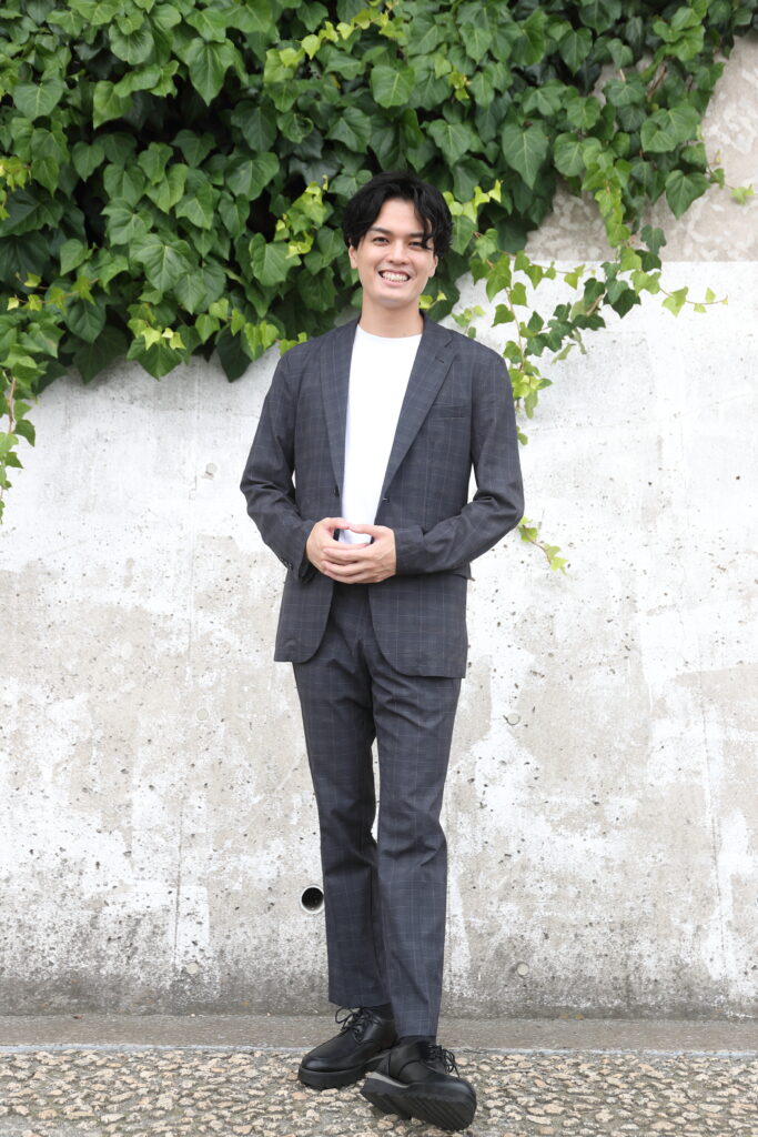 グレーのスーツを着た男性が緑の壁の前で笑顔で立っている